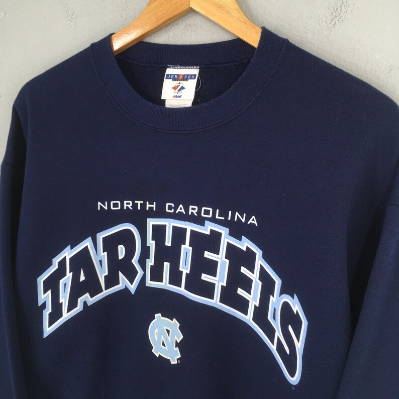 May include: Navy blue sweatshirt with the words "NORTH CAROLINA" above "TAR HEELS" in a light blue, outlined font. A light blue logo is below the text. The sweatshirt is hanging on a wooden hanger.