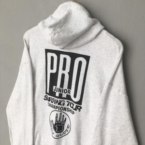 Puede incluir: Una sudadera con capucha gris claro con una capucha y un gran gr&aacute;fico negro en la espalda. El gr&aacute;fico dice "PRO JUNIOR SPRING TOUR CHAMPIONSHIP" con un logotipo de mano. La sudadera con capucha est&aacute; hecha de un material suave y c&oacute;modo.