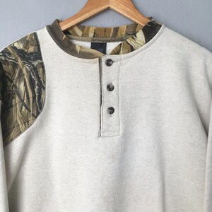 May include: A light gray sweatshirt with a camouflage pattern on the left shoulder and collar. The shirt has a Henley neckline with three dark buttons. The collar and shoulder camouflage pattern features shades of brown, green, and tan.
