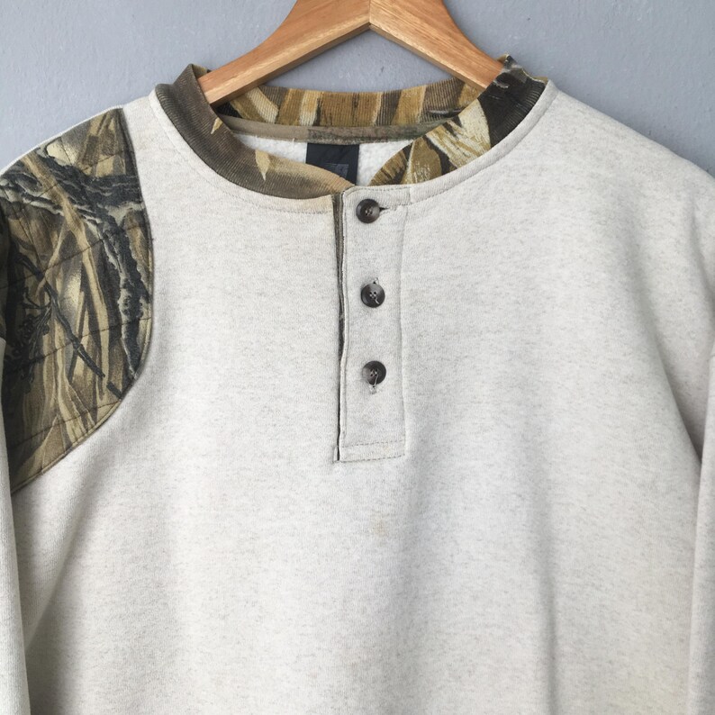 May include: A light gray sweatshirt with a camouflage pattern on the left shoulder and collar. The shirt has a Henley neckline with three dark buttons. The collar and shoulder camouflage pattern features shades of brown, green, and tan.