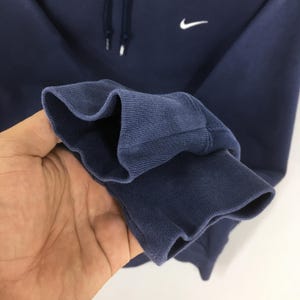 Pu&ograve; includere: Felpa con cappuccio blu navy con logo Nike bianco. La felpa ha un cappuccio con coulisse e polsini a coste. Primo piano del polsino.