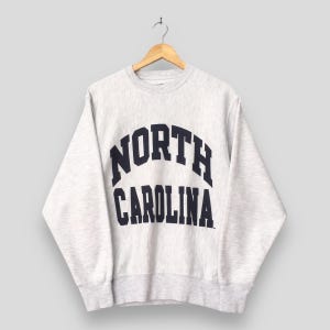 May include: A light gray sweatshirt with the words "NORTH CAROLINA" in navy blue arched lettering. The sweatshirt is hanging on a wooden hanger against a light gray background. The sweatshirt has a crew neck and long sleeves.