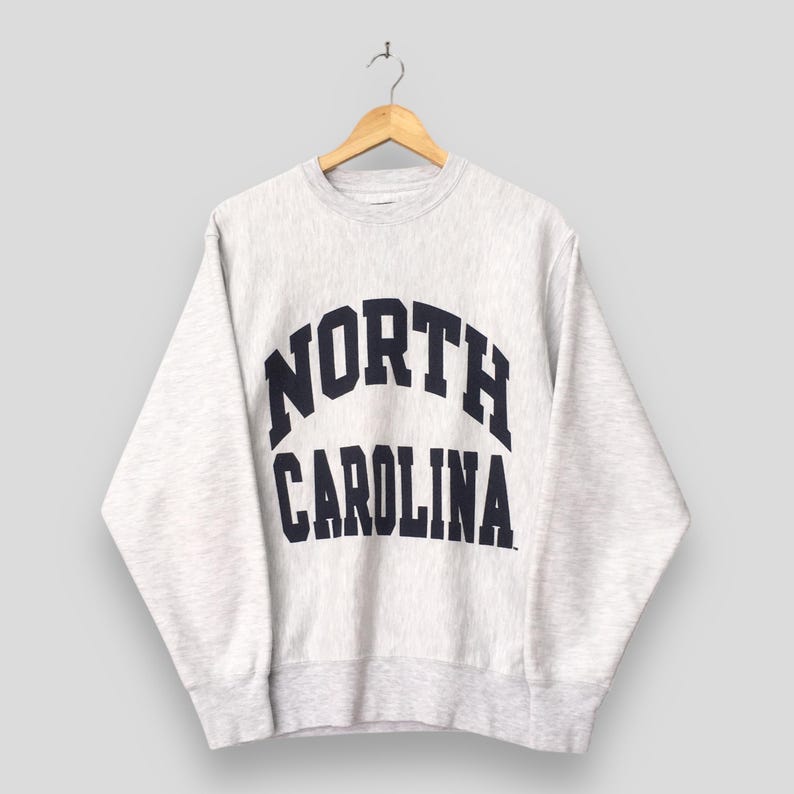May include: A light gray sweatshirt with the words "NORTH CAROLINA" in navy blue arched lettering. The sweatshirt is hanging on a wooden hanger against a light gray background. The sweatshirt has a crew neck and long sleeves.