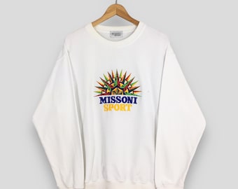 Puede incluir: Sudadera blanca de cuello redondo con un diseño geométrico colorido y el texto "MISSONI SPORT" en azul marino y amarillo. La sudadera está colgada en una percha de madera.