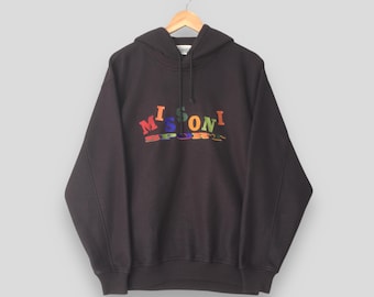 Puede incluir: Una sudadera con capucha negra con la palabra "MISSONI SPORT" en letras coloridas en el pecho. La sudadera está colgada de una percha de madera sobre un fondo claro. Las letras son rojas, verdes, azules y naranjas.