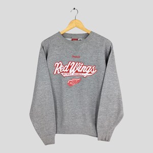 May include: A gray Reebok sweatshirt featuring the Detroit Red Wings logo in red and white. The logo includes the words "Red Wings" in a script font, the team's winged wheel logo, and the words "Detroit Hockey".