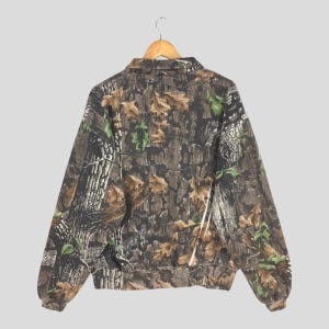 K&ouml;nnte beinhalten: Eine Tarnjacke mit durchgehendem Rei&szlig;verschluss und Kragen, die an einem Holzb&uuml;gel h&auml;ngt. Das Muster der Jacke ahmt Baumrinde, Bl&auml;tter und &Auml;ste in Braun-, Gr&uuml;n- und Beiget&ouml;nen nach. Die R&uuml;ckansicht der Jacke.