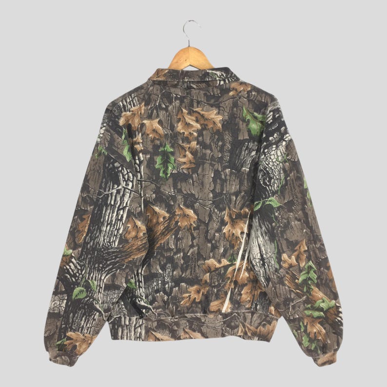 K&ouml;nnte beinhalten: Eine Tarnjacke mit durchgehendem Rei&szlig;verschluss und Kragen, die an einem Holzb&uuml;gel h&auml;ngt. Das Muster der Jacke ahmt Baumrinde, Bl&auml;tter und &Auml;ste in Braun-, Gr&uuml;n- und Beiget&ouml;nen nach. Die R&uuml;ckansicht der Jacke.