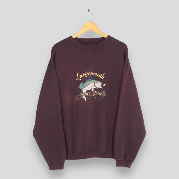 Könnte beinhalten: Ein weinrotes Sweatshirt mit einer Grafik eines Großmaulbasses. Das Design enthält die Worte "Largemouth Legends Gamefish" in einer stilisierten Schriftart. Das Sweatshirt hängt an einem Holzkleiderbügel.