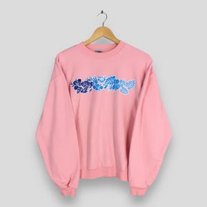 May include: A pale pink sweatshirt with a crew neck, suspended from a wooden hanger. The front displays a floral design in shades of blue, with the word "ROXY" above the floral design.