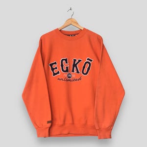 May include: An orange long-sleeve crewneck sweatshirt featuring the text "ECKO" in large dark blue letters with white outlines, and the text "unlimited" in smaller cursive script below, with the number "1972" and a small circular logo.