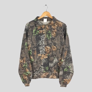 K&ouml;nnte beinhalten: Ein Sweatshirt mit Camouflage-Muster und einem Viertel-Rei&szlig;verschluss. Das Design zeigt eine Mischung aus braunen, gr&uuml;nen und beigen Farben, die eine Waldumgebung nachahmen. Das lang&auml;rmlige Oberteil h&auml;ngt an einem Holzb&uuml;gel.