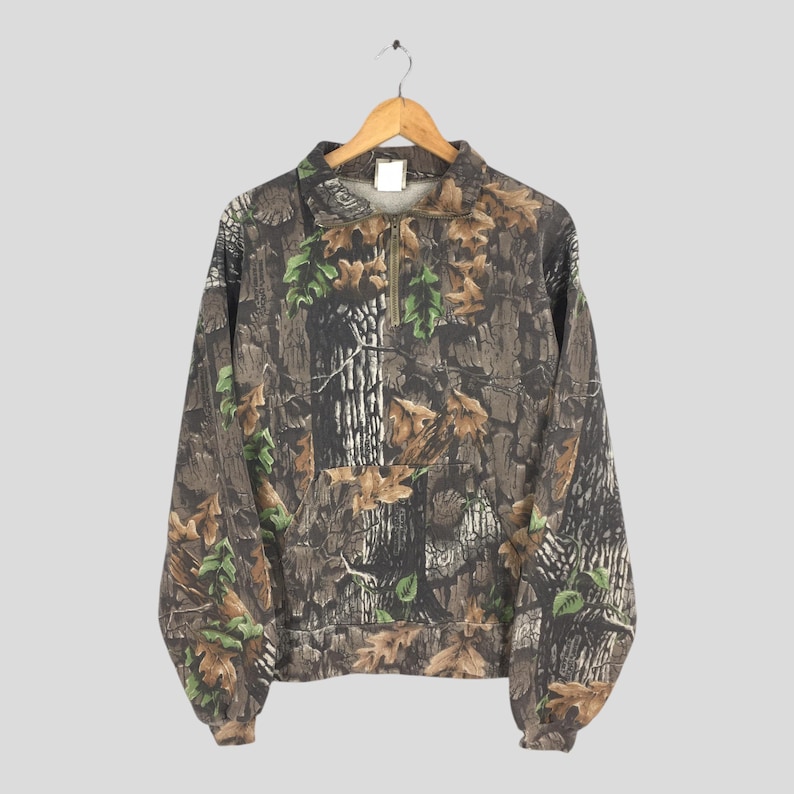 K&ouml;nnte beinhalten: Ein Sweatshirt mit Camouflage-Muster und einem Viertel-Rei&szlig;verschluss. Das Design zeigt eine Mischung aus braunen, gr&uuml;nen und beigen Farben, die eine Waldumgebung nachahmen. Das lang&auml;rmlige Oberteil h&auml;ngt an einem Holzb&uuml;gel.