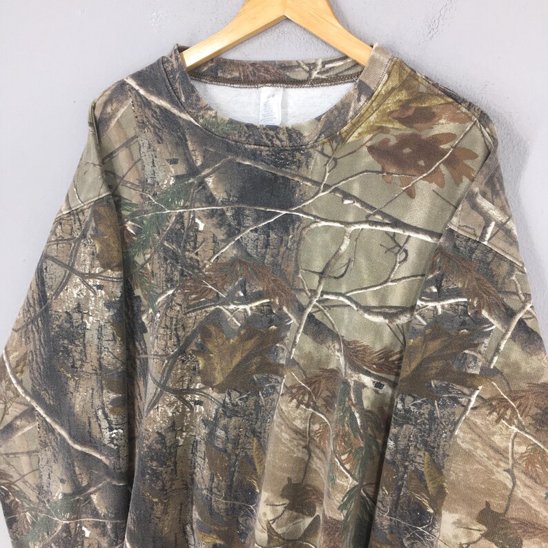 May include: A crew neck sweatshirt with a camouflage pattern in shades of brown, green, and tan. The sweatshirt is hanging on a wooden hanger. The fabric has a soft, slightly worn appearance.
