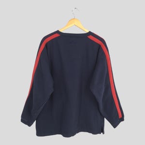 Puede incluir: Sudadera azul marino con rayas rojas en las mangas. La parte superior de manga larga est&aacute; colgada de una percha de madera. La sudadera tiene cuello redondo y un corte holgado.