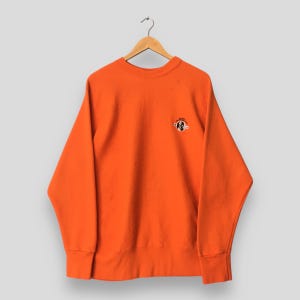 May include: Bright orange sweatshirt with a small embroidered design on the upper left chest. The sweatshirt has a crew neck and ribbed cuffs and hem. The design is a black and white cartoon character.