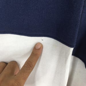 May include: Close-up of a navy blue and white color-blocked sweatshirt. The fabric is divided horizontally, with navy blue at the top and white at the bottom. A finger points to a small hole in the white section.