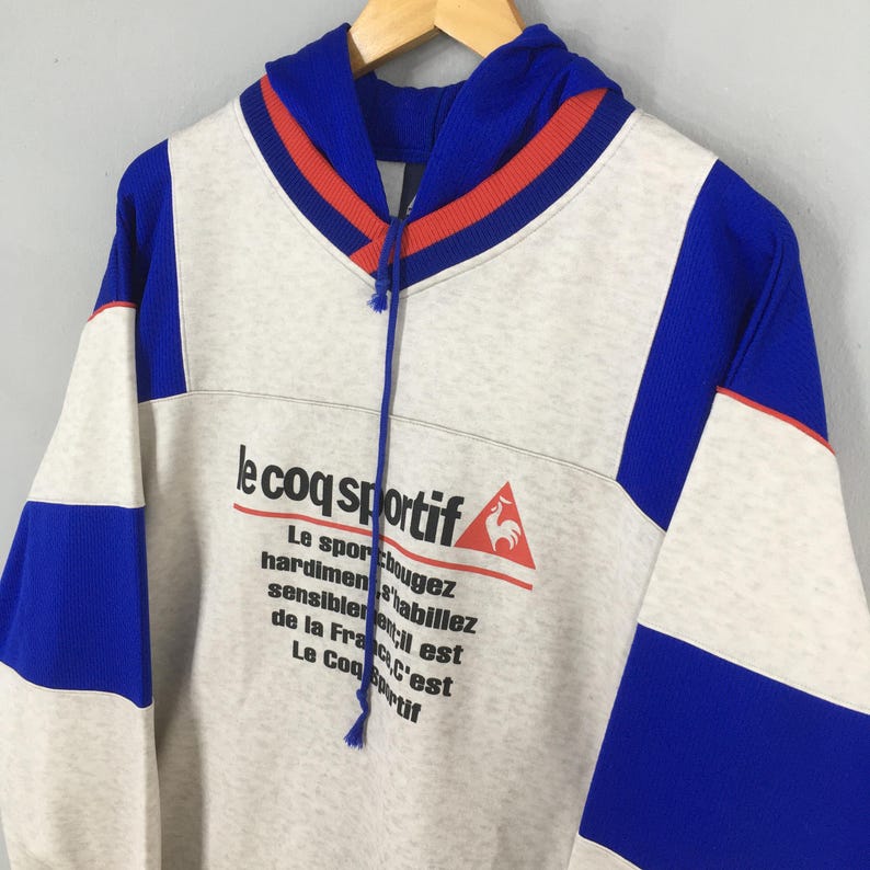May include: A gray, blue, and white Le Coq Sportif hoodie with a drawstring hood. The hoodie features blue sleeves and accents, with the brand's logo and text printed on the front. The text on the front reads "Le sport bougez, hardiment habillez, sensiblement est, de la France, C'est Le Coq sportif."