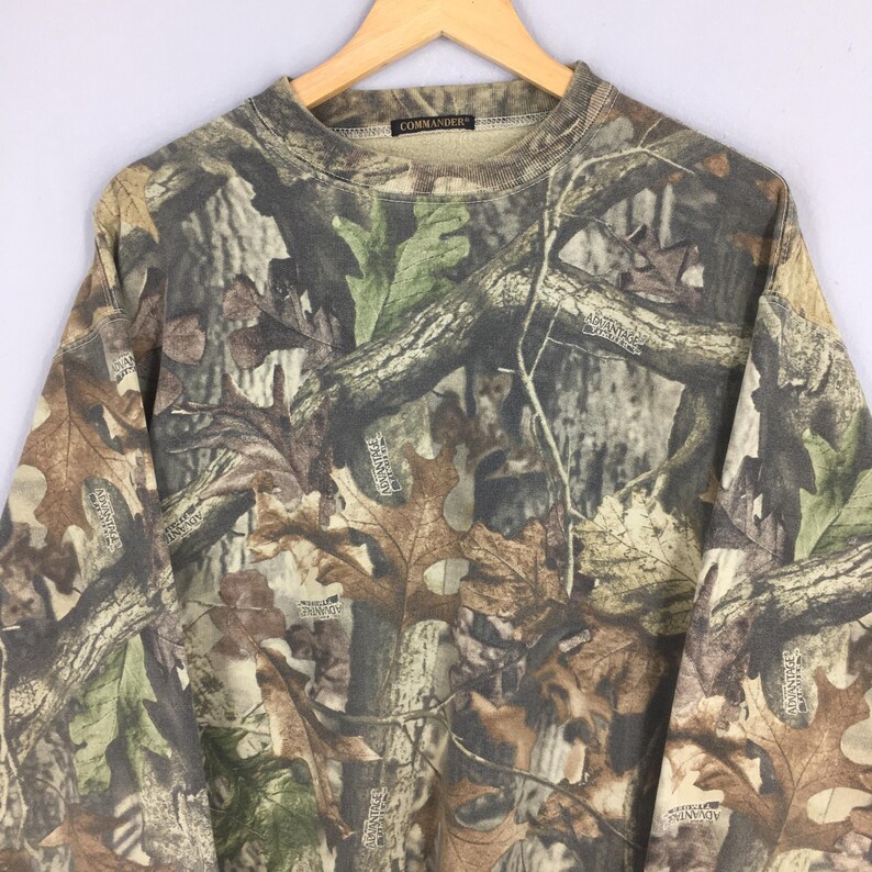 K&ouml;nnte beinhalten: Ein Sweatshirt mit Camouflage-Muster, Rundhalsausschnitt und langen &Auml;rmeln. Das Design zeigt eine Mischung aus Braun-, Gr&uuml;n- und Beiget&ouml;nen, die eine Waldumgebung nachahmen. Das Wort "COMMANDER" ist am Kragen sichtbar.