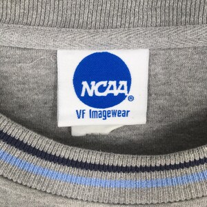 Puede incluir: Primer plano de una sudadera gris de cuello redondo con una etiqueta blanca con el logotipo azul de la NCAA y el texto "VF Imagewear". El cuello tiene rayas azul marino y celeste.