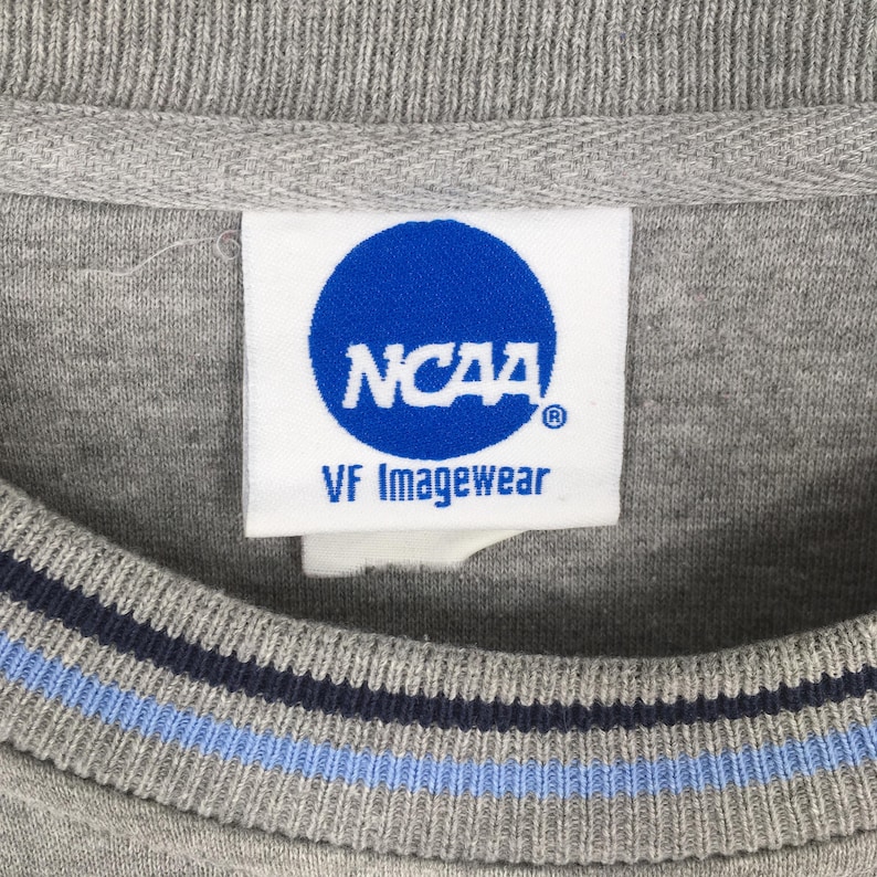 Puede incluir: Primer plano de una sudadera gris de cuello redondo con una etiqueta blanca con el logotipo azul de la NCAA y el texto "VF Imagewear". El cuello tiene rayas azul marino y celeste.