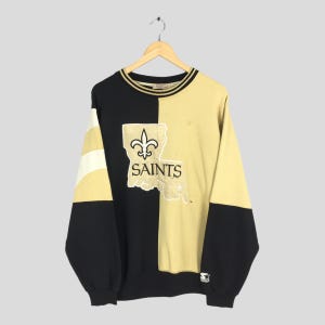 May include: Black and gold Saints sweatshirt with a split design. The left side is black with white and gold stripes on the sleeve. The right side is gold with the Saints logo and the word "SAINTS" in white.
