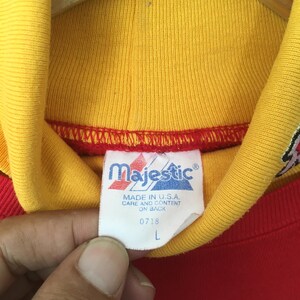 Peut inclure: Un v&ecirc;tement rouge et jaune avec une &eacute;tiquette de marque Majestic blanche au niveau du col. L'&eacute;tiquette affiche le texte Majestic, MADE IN U.S.A., CARE AND CONTENT ON BACK, 0718, et la taille L. Le col est jaune avec une bordure rouge.