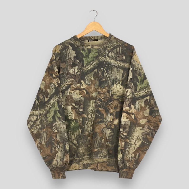 K&ouml;nnte beinhalten: Ein lang&auml;rmeliges Sweatshirt mit Camouflage-Muster h&auml;ngt an einem Holzb&uuml;gel. Das Muster zeigt Braunt&ouml;ne, Gr&uuml;n und Beige, die einer Waldszene &auml;hneln. Das Sweatshirt hat einen Rundhalsausschnitt und gerippte B&uuml;ndchen und Saum.