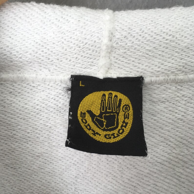 Puede incluir: Primer plano de una sudadera con capucha blanca con una etiqueta negra. La etiqueta presenta un c&iacute;rculo amarillo con un gr&aacute;fico de mano y las palabras "BODY GLOVE" en amarillo. La etiqueta tambi&eacute;n tiene la letra "L" en blanco.