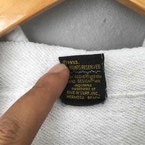 Puede incluir: Primer plano de una etiqueta negra con texto blanco. La etiqueta dice "&copy; 1953, TODOS LOS DERECHOS RESERVADOS" y menciona "DIVE N'SURF, INC., HERMOSA BEACH". La etiqueta est&aacute; adherida a una tela blanca.
