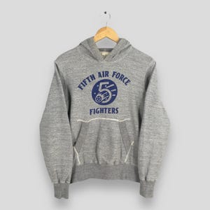 May include: Gray hooded sweatshirt with a front pocket and ribbed cuffs. The sweatshirt features a blue and white graphic that reads "FIFTH AIR FORCE FIGHTERS". The hoodie is hanging on a wooden hanger.
