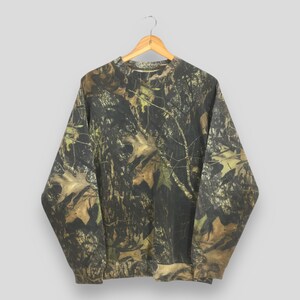 May include: A long-sleeved camouflage sweatshirt in shades of brown, green, and black, hanging on a wooden hanger. The pattern mimics a forest environment, with leaf and branch details.