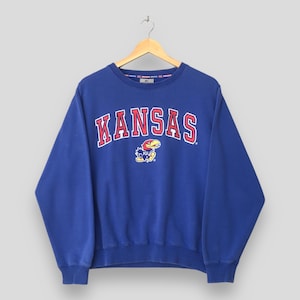 May include: A royal blue sweatshirt featuring the word "KANSAS" in red and white lettering. Below the text is a graphic of a Jayhawk mascot. The sweatshirt has a crew neck and long sleeves.