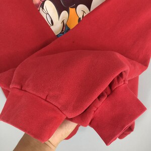 May include: A red sweatshirt featuring a graphic of Minnie Mouse. The sweatshirt has long sleeves with ribbed cuffs. The graphic is in the center of the chest. The fabric appears to be a soft, comfortable material.