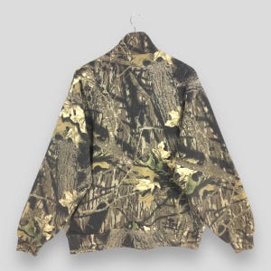 Pode incluir: Casaco camuflado com z&iacute;per e gola alta, com um padr&atilde;o de casca de &aacute;rvore e folhas marrons, verdes e bege. O casaco est&aacute; pendurado em um cabide. Possui bainha e punhos canelados.