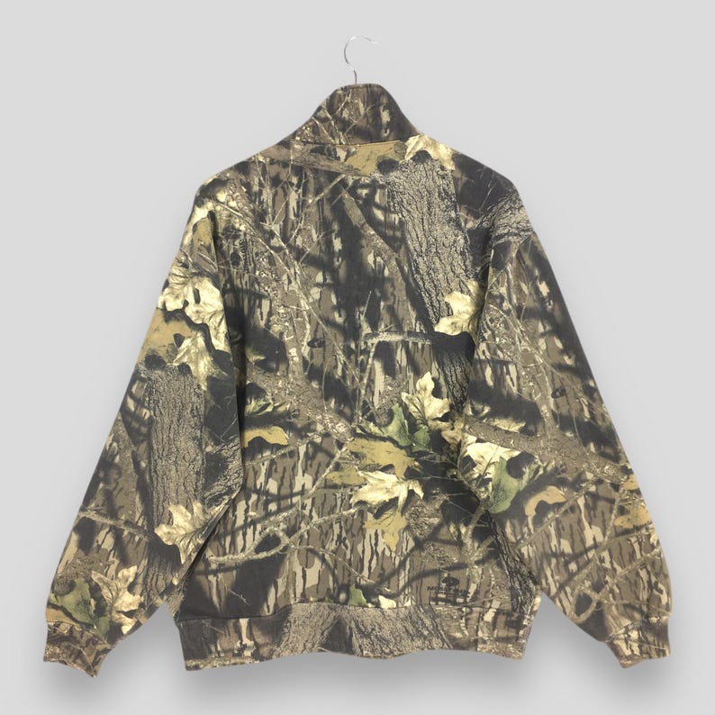 Pode incluir: Casaco camuflado com z&iacute;per e gola alta, com um padr&atilde;o de casca de &aacute;rvore e folhas marrons, verdes e bege. O casaco est&aacute; pendurado em um cabide. Possui bainha e punhos canelados.