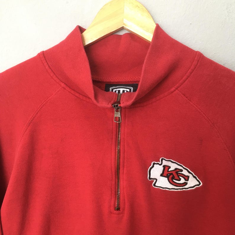 Op de afbeelding: Een rood sweatshirt met een korte rits en een opstaande kraag. Op de linkerborst bevindt zich een geborduurde patch in de vorm van een pijlpunt met een witte rand en de rode letters KC in het midden van het ontwerp.