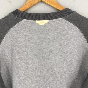 K&ouml;nnte beinhalten: Nahaufnahme eines grauen Sweatshirts mit dunkelgrauen Raglan&auml;rmeln. Der Halsausschnitt ist dunkelgrau und ein kleines gelbes Etikett mit einem Logo ist auf der R&uuml;ckseite angen&auml;ht. Das Sweatshirt h&auml;ngt an einem Holzb&uuml;gel.