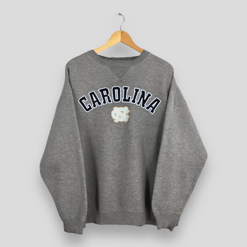 May include: A gray sweatshirt with the word "CAROLINA" arched across the chest in white lettering with a navy blue outline. Below the word is a light blue and white emblem. The sweatshirt is hanging on a wooden hanger.
