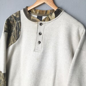 May include: A light gray sweatshirt with a camouflage pattern on the shoulders and collar. The long-sleeved shirt has a three-button placket and is hanging on a wooden hanger.