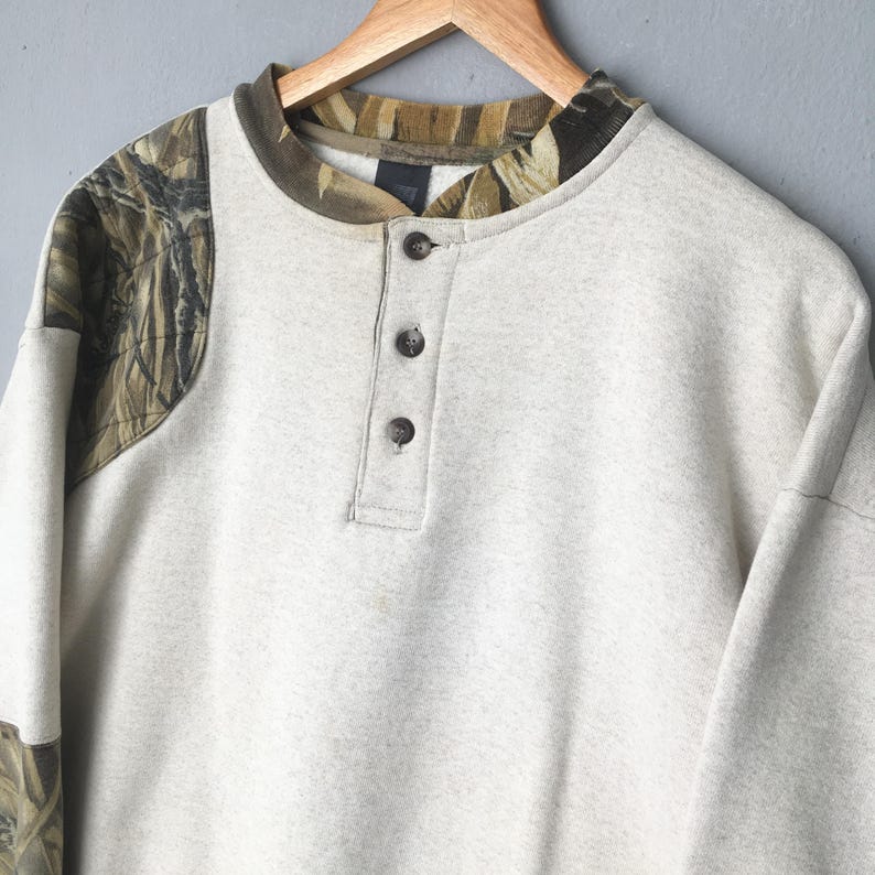May include: A light gray sweatshirt with a camouflage pattern on the shoulders and collar. The long-sleeved shirt has a three-button placket and is hanging on a wooden hanger.