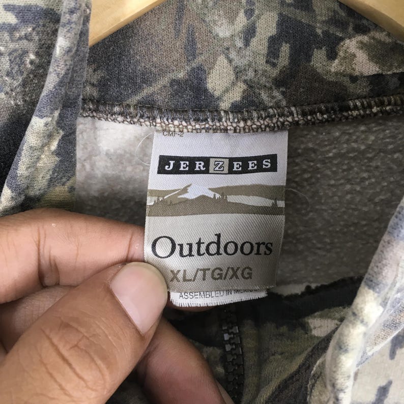 May include: Close-up of a camouflage-patterned sweatshirt with the tag "JERZEES Outdoors" visible. The tag also indicates the size as XL/TG/XG. The sweatshirt has a zipper and is made of a soft material.