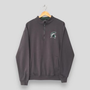 Puede incluir: Una sudadera gris carbón con cremallera de un cuarto con un logotipo de casco espartano en verde y blanco en el pecho izquierdo. La sudadera tiene un pequeño bolsillo en la parte inferior delantera y un forro verde dentro del cuello.