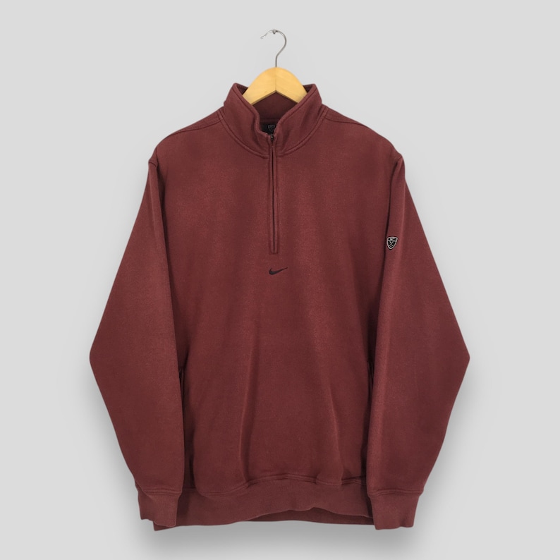 May include: A maroon Nike pullover sweatshirt with a quarter-zip fastening and a small black Nike logo on the chest. The sweatshirt has a high collar and a small Nike logo on the left sleeve. The garment is hanging on a wooden hanger.