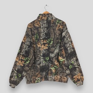 May include: A full-zip camouflage jacket with a high collar, featuring a pattern of brown tree bark, green leaves, and tan foliage. The jacket is hanging on a wooden hanger against a neutral background.