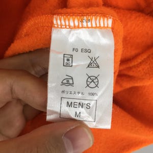 May include: Close-up of an orange fleece garment tag. The tag displays care symbols, the text "F0 ESQ", and "MEN'S M". The fabric is 100% polyester.