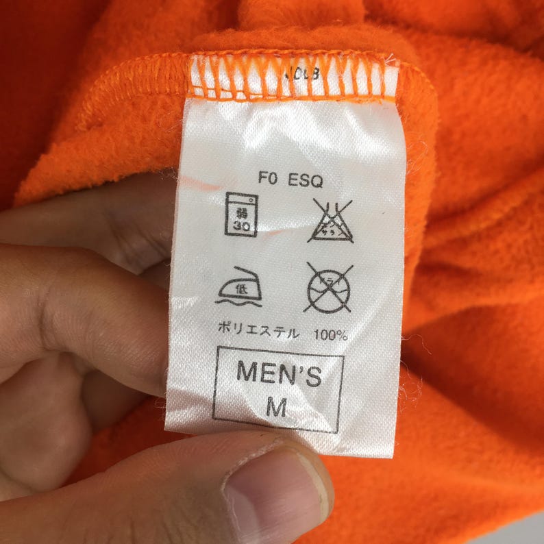 May include: Close-up of an orange fleece garment tag. The tag displays care symbols, the text "F0 ESQ", and "MEN'S M". The fabric is 100% polyester.