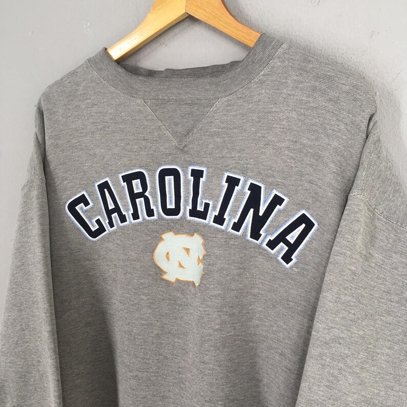 May include: A gray sweatshirt with the word "CAROLINA" in navy blue letters with a white outline. Below the word is a light blue and gold logo. The sweatshirt is hanging on a wooden hanger.