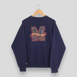 Puede incluir: Sudadera azul marino con un gráfico floral de la letra "M" y las palabras "Morehead State" en una pancarta. La sudadera está colgada en una percha de madera. La sudadera es de cuello redondo.