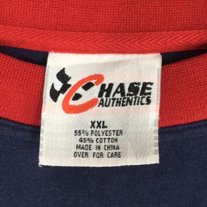 May include: Close-up of a navy blue and red shirt with a tag. The tag reads "CHASE AUTHENTICS" with a logo. The tag also indicates the shirt is XXL, made of 55% polyester and 45% cotton, and made in China.